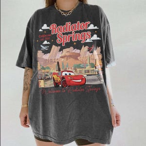 May include: A white ceramic mug with a black handle and the text "But First, Coffee" in a black, handwritten-style font. The mug is on a white surface.