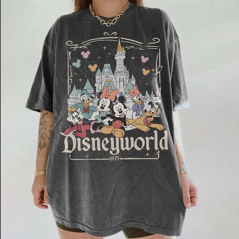 May include: Dark gray t-shirt featuring a Disneyworld graphic. The design includes Mickey and Minnie Mouse, Donald and Daisy Duck, and Goofy in front of a castle. The text "Disneyworld" and "1971" are also visible.