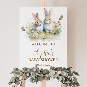 Op de afbeelding: Een welkomstbord voor een babyshower met aquarel illustraties van twee konijnen, vlinders en bloemen. Het bord leest "Welkom bij Sophia's Baby Shower" met een datum. Het bord wordt weergegeven op een houten ezel.