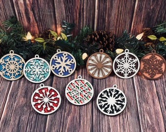 May include: A collection of nine circular wooden ornaments featuring intricate snowflake cutouts. The designs include various backings such as solid blue, red glitter, teal glitter, black glitter, striped patterns, and natural wood finishes.
