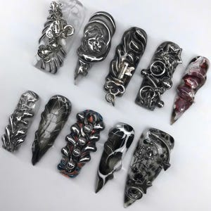 K&ouml;nnte beinhalten: Eine Auswahl an modellierten Nagelkunst-Designs. Die Kollektion umfasst verschiedene Formen und Farben, darunter transparent, schwarz, silber und rot. Einige N&auml;gel haben aufwendige Metallverzierungen, w&auml;hrend andere Marmormuster aufweisen.