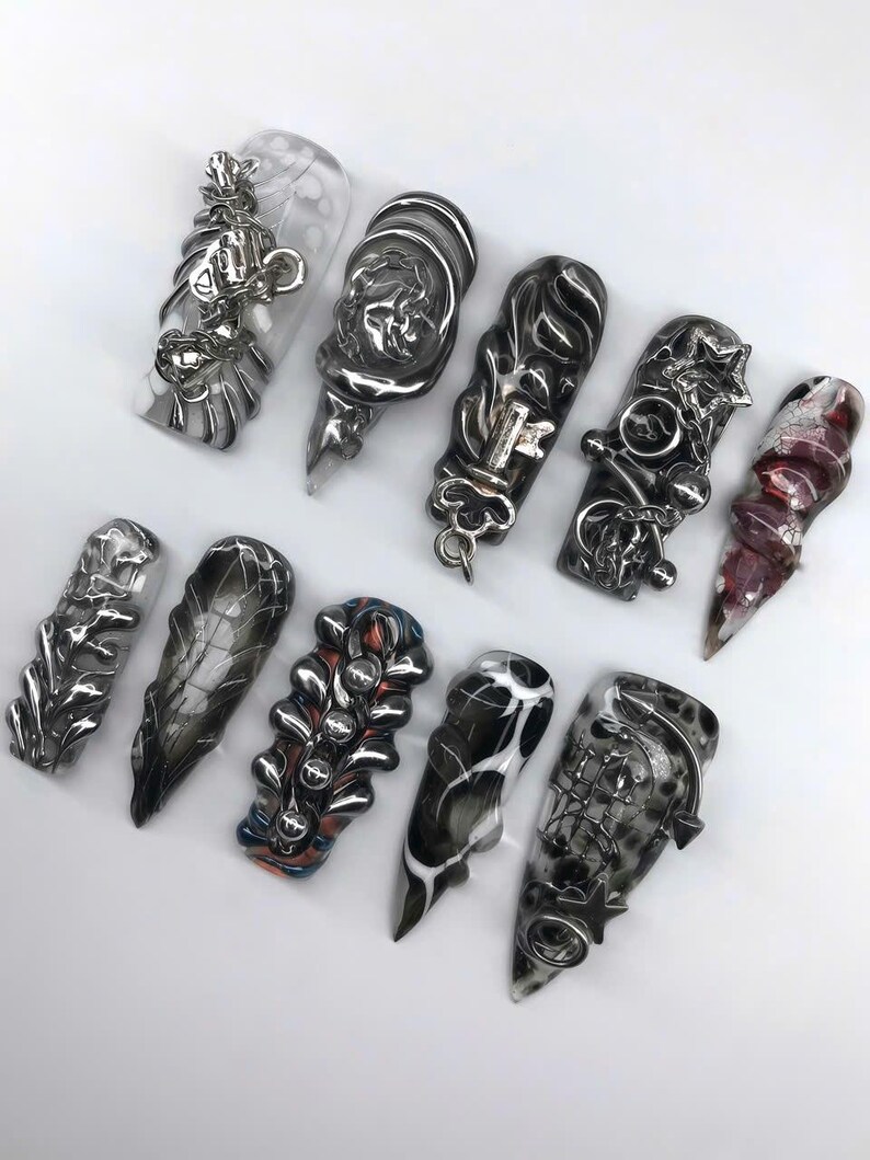 K&ouml;nnte beinhalten: Eine Auswahl an modellierten Nagelkunst-Designs. Die Kollektion umfasst verschiedene Formen und Farben, darunter transparent, schwarz, silber und rot. Einige N&auml;gel haben aufwendige Metallverzierungen, w&auml;hrend andere Marmormuster aufweisen.