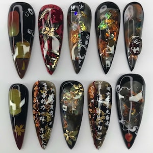 May include: A set of ten long, almond-shaped artificial nails. Each nail features a unique design with a glossy finish. Designs include gold leaf accents, Japanese calligraphy, and abstract patterns in shades of black, red, and brown.
