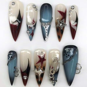 Pode incluir: Um conjunto de dez unhas artificiais longas e pontiagudas com vários designs. As unhas apresentam uma mistura de cores, incluindo branco, preto, vermelho e azul, com enfeites como tachas prateadas, correntes e berloques de estrela. Algumas unhas têm efeitos gradientes.