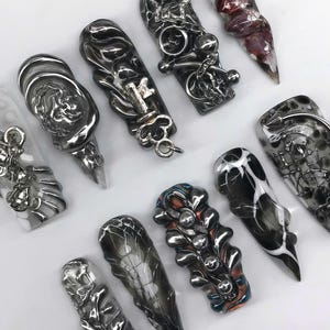 K&ouml;nnte beinhalten: Eine Auswahl an einzigartig gestalteter Nagelkunst mit verschiedenen Formen und Verzierungen. Die N&auml;gel zeigen eine Reihe von Farben, darunter Silber, Schwarz und Rot, mit komplizierten Mustern und Texturen. Einige N&auml;gel haben Stern-, Schl&uuml;ssel- und Ketten Details.