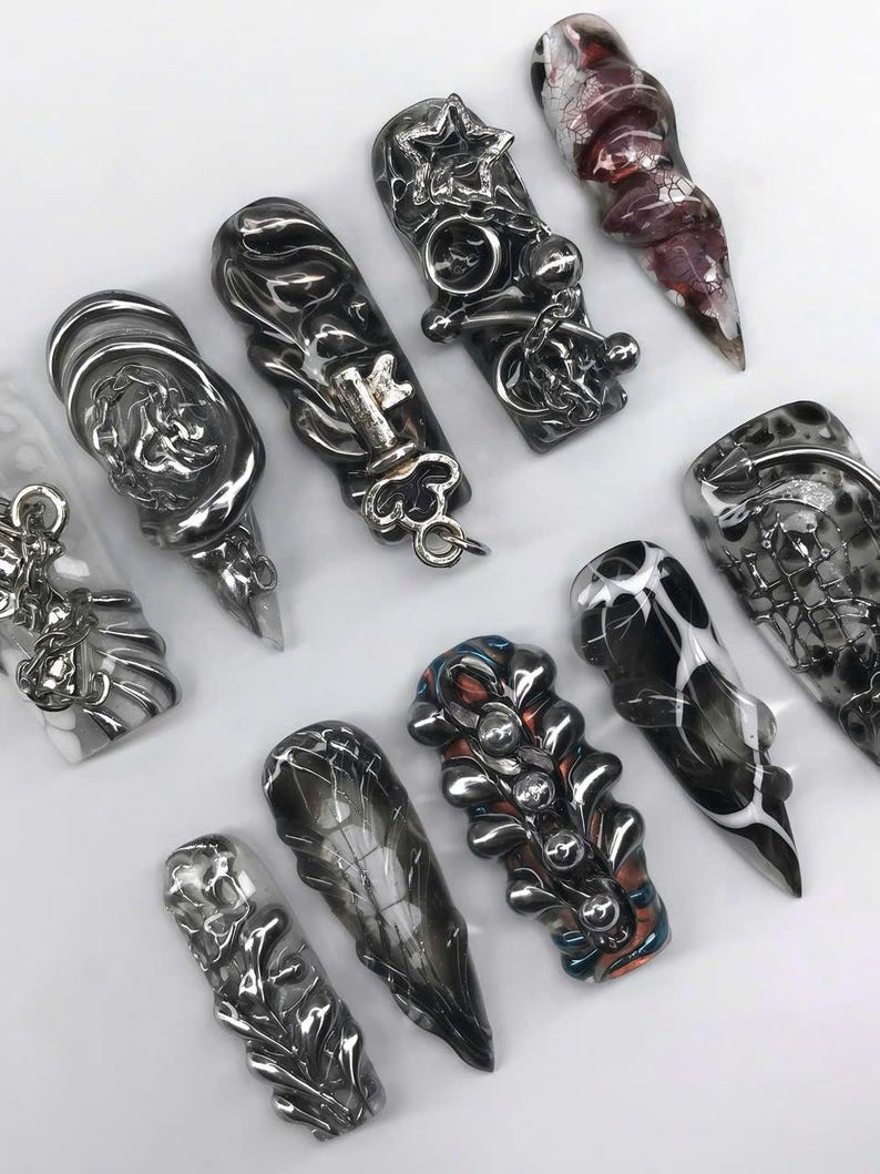 K&ouml;nnte beinhalten: Eine Auswahl an einzigartig gestalteter Nagelkunst mit verschiedenen Formen und Verzierungen. Die N&auml;gel zeigen eine Reihe von Farben, darunter Silber, Schwarz und Rot, mit komplizierten Mustern und Texturen. Einige N&auml;gel haben Stern-, Schl&uuml;ssel- und Ketten Details.