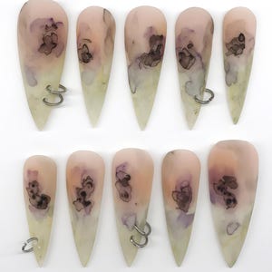 Puede incluir: Un conjunto de diez uñas postizas en forma de almendra con una base de color melocotón pálido y crema, con manchas abstractas de tinta en color morado oscuro y negro. Tres de las uñas incluyen pequeños anillos metálicos plateados.