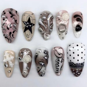 May include: Ten artificial nails with diverse designs. The nails showcase beige, brown, and white hues, adorned with stars, swirls, and rhinestones. Some nails feature text, while others have lace or floral details. A variety of nail art styles.