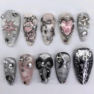 May include: A collection of ten artificial nails with diverse designs. The nails showcase silver, pink, and black embellishments, including stars, hearts, bows, and a Hello Kitty design. Some nails have the text "miu miu".