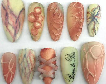 May include: A set of ten almond-shaped press-on nails featuring various designs in shades of peach, cream, and red. The nails display textures including raised circular bumps, vein-like patterns, crisscross bands, and the text Born to Die.