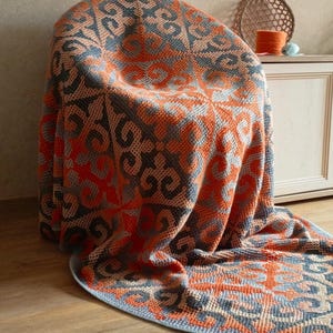 Op de afbeelding: Een gehaakte deken met een geometrisch patroon in oranje, grijs en beige, gedrapeerd over een stoel. Het ontwerp van de deken is zichtbaar, met de tekst "CROCHETED BY NADEZHDA GOLIKOVA" onderaan.