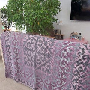 May include: A crocheted blanket in shades of purple and gray, draped over a sofa. The blanket features a repeating geometric pattern. The text at the bottom reads "Made from this pattern Crocheted by Tatiana Seredkina."