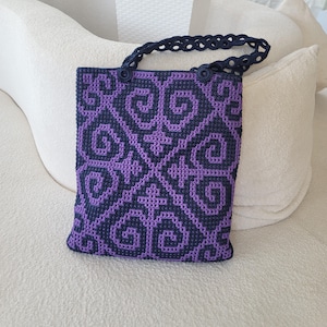 Puede incluir: Un bolso rectangular de ganchillo con un patrón geométrico en azul marino oscuro y morado vibrante. El bolso tiene un diseño plano y estructurado con un asa de ganchillo a juego en azul marino que presenta un motivo repetitivo de círculos calados.