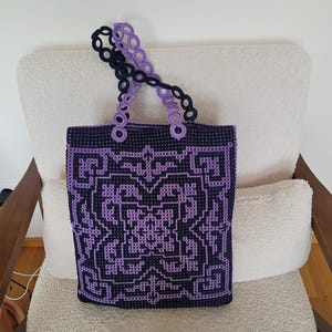 May include: A rectangular crochet tote bag featuring a dark navy blue and light purple geometric pattern. The bag has two handles constructed from a series of interconnected circular crochet rings, alternating between navy blue and light purple colors.