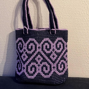 May include: A navy blue crochet tote bag with a lavender geometric design. The bag has matching navy handles and trim. The textured crochet pattern is visible on the bag's surface. Ideal for carrying everyday items.