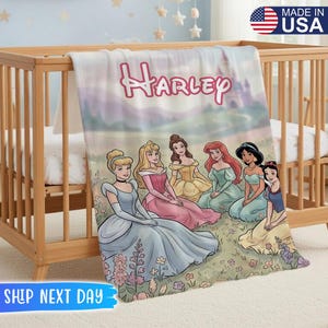 May include: A personalized blanket featuring the name "Harley" in pink script above a scene with Disney princesses. The blanket has a soft, pastel color scheme and is draped over a wooden crib. The words "MADE IN USA" are visible.