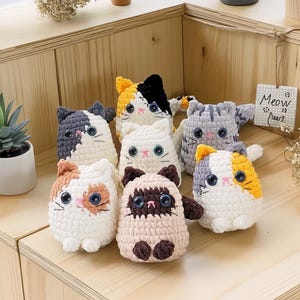 Könnte beinhalten: Eine Sammlung handgefertigter Häkelkatzen-Plüschtiere in verschiedenen Farben und Mustern, darunter Calico-, Grautabby- und Siam-Designs. Jedes Plüschtier hat große, ausdrucksstarke Augen und eine niedliche, abgerundete Form. Ein kleines Schild sagt "Meow".