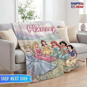 May include: A soft, personalized blanket featuring Disney princesses, including Cinderella, Aurora, Belle, Ariel, Jasmine, and Snow White. The name "Harley" is displayed in pink script across the top. The blanket is draped over a gray sofa.