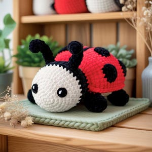Könnte beinhalten: Ein handgefertigtes Häkel-Marienkäfer-Spielzeug. Der Marienkäfer hat einen weißen Kopf mit schwarzen Augen und Fühlern. Sein Körper ist rot mit schwarzen Punkten. Es liegt auf einer grünen Strickmatte, mit einem Holzregal und Topfpflanzen im Hintergrund.