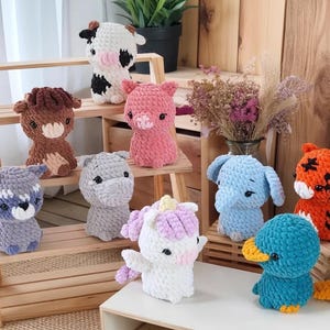 Könnte beinhalten: Eine Sammlung handgefertigter gehäkelter Tierplüschtiere, darunter eine Kuh, ein Schwein, ein Pferd, ein Waschbär, ein Nilpferd, ein Einhorn, ein Elefant, ein Tiger und eine Ente. Die Plüschtiere sind in verschiedenen Farben und Größen auf Holzregalen und einer weißen Oberfläche ausgestellt.