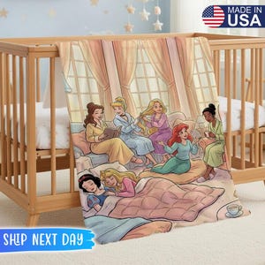 以下が含まれることがあります： 柔らかいピンクとブルーの毛布。ディズニーのプリンセスが描かれた漫画のイラストが特徴です。木製のベビーベッドに掛けられており、「MADE IN USA」の文字が見えます。