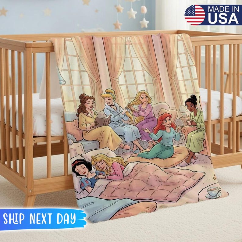 以下が含まれることがあります： 柔らかいピンクとブルーの毛布。ディズニーのプリンセスが描かれた漫画のイラストが特徴です。木製のベビーベッドに掛けられており、「MADE IN USA」の文字が見えます。