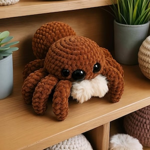 Könnte beinhalten: Eine handgefertigte braune Häkelspinne mit großen schwarzen Augen und einem flauschigen weißen Mund. Das Plüschtier hat acht Beine und einen runden Körper. Die Spinne wird auf einem Holzregal mit anderen dekorativen Gegenständen ausgestellt.