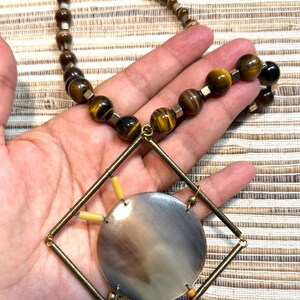 Pode incluir: Um colar artesanal com contas castanhas e douradas, incluindo pedras olho de tigre. O pingente tem um design geométrico com um grande centro redondo de concha iridescente. O colar é segurado em uma mão.