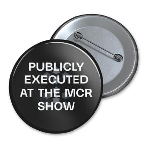 May include: Two round black buttons with silver pin backs. The front button has white text that reads "PUBLICLY EXECUTED AT THE MCR SHOW". The second button is partially visible, showing the silver pin back.