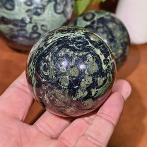 K&ouml;nnte beinhalten: Eine polierte Kugel aus gr&uuml;nem und schwarzem Stein, gehalten in einer Hand. Der Orb hat ein wirbelndes Muster aus dunkelgr&uuml;n und schwarz, mit helleren gr&uuml;nen Flecken. Im Hintergrund sind weitere &auml;hnliche Kugeln zu sehen, was auf eine Sammlung von Dekorationsartikeln hindeutet.