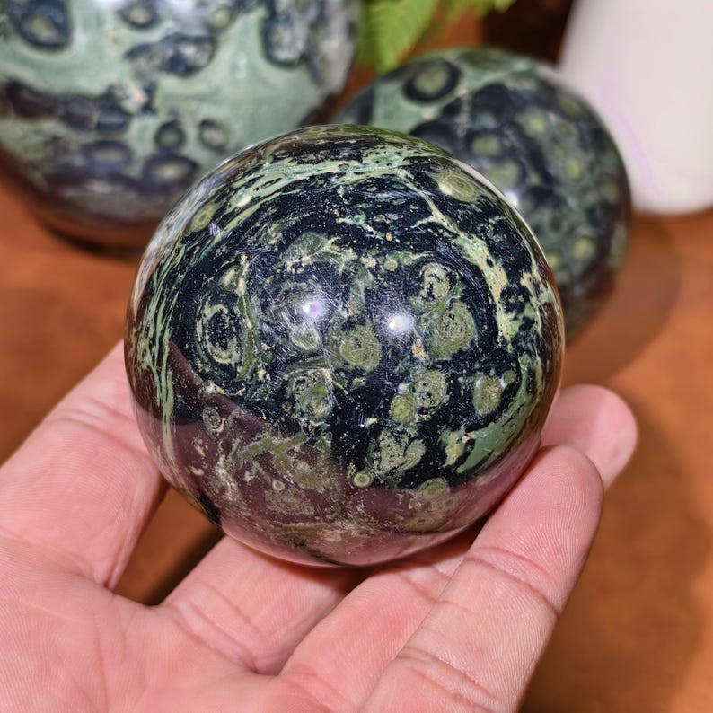 K&ouml;nnte beinhalten: Eine polierte Kugel aus gr&uuml;nem und schwarzem Stein, gehalten in einer Hand. Der Orb hat ein wirbelndes Muster aus dunkelgr&uuml;n und schwarz, mit helleren gr&uuml;nen Flecken. Im Hintergrund sind weitere &auml;hnliche Kugeln zu sehen, was auf eine Sammlung von Dekorationsartikeln hindeutet.