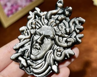 May include: A detailed, metallic Medusa head pendant. The pendant depicts a face with an open mouth and snake-like hair. The colour is a dark, antique silver. The design is highly detailed and intricate.