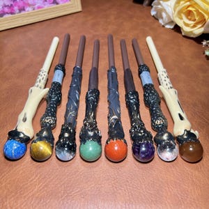 May include: A collection of eight fantasy wands with various designs. Each wand features a dark brown handle, ornate black detailing, and a gemstone at the base. Gemstones include blue, yellow, clear, green, red, purple, clear, and brown.