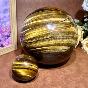以下が含まれることがあります： 研磨されたタイガーアイの球が2つ。 大きな球の直径は約15cm、小さな球の直径は約5cmです。 球は、明るい色合いと暗い色合いの帯を持つ、豊かな金茶色をしています。