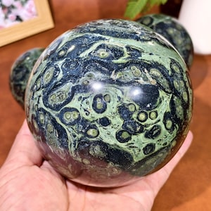May include: A polished, spherical gemstone with a unique pattern of dark green, light green, and black swirls and spots. The sphere is held in a hand, showcasing its smooth, reflective surface. The gemstone's intricate design is the focal point.