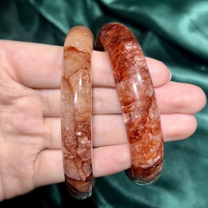 Op de afbeelding: Twee heldere, roodbruine kristallen armbanden. De armbanden hebben een gepolijst, glad oppervlak met natuurlijke insluitsels die een uniek patroon creëren. De kleur varieert van licht- tot donkerbruin, met een doorschijnend uiterlijk. De armbanden hebben een diameter van ongeveer 6,35 cm.