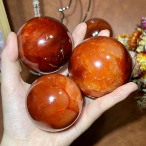 Può includere: Quattro sfere di corniola lucidata di varie dimensioni. Le sfere variano di colore dal rosso intenso all'arancione, con motivi naturali visibili sulle loro superfici. Le sfere sono tenute in una mano, mostrando le loro qualità lisce e riflettenti.