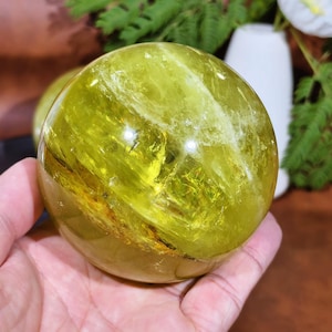 Puede incluir: Una gran esfera de cristal translúcido de color amarillo verdoso. La esfera tiene patrones e inclusiones internas, con una superficie pulida y reflectante. La bola de cristal se sostiene en una mano, con un fondo borroso de vegetación y un jarrón blanco.