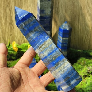Può includere: Una collezione di obelischi in lapislazzuli blu. Gli obelischi hanno una finitura lucida, che mostra il colore blu naturale con venature bianche e dorate. Gli obelischi sono su un letto di muschio verde.