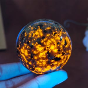 Puede incluir: Una esfera brillante con un patrón moteado de negro y naranja. El orbe parece estar hecho de un material translúcido, con la luz naranja que emana del interior. La esfera se sostiene en una mano, con una luz azul que la ilumina.
