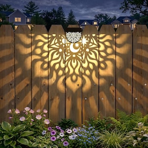 May include: A solar-powered fence light with a decorative, laser-cut design. The light casts a pattern of intricate shapes onto the wooden fence, featuring a crescent moon, star, and floral elements. The warm glow illuminates the surrounding garden and flowers.
