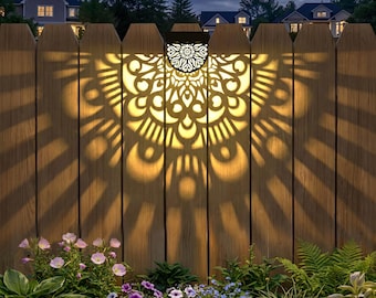 May include: A solar-powered fence light with a decorative, openwork design. The light casts a mandala-like pattern on the wooden fence. The image shows the light illuminating the fence at night, with flowers and foliage in the foreground.