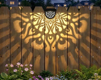 May include: A solar-powered outdoor light fixture mounted on a wooden fence. The black fixture casts a decorative floral pattern onto the fence. The fence is surrounded by a garden with various flowers and greenery, with a night sky background.