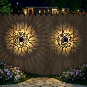 May include: Two solar-powered sunflower-shaped lights mounted on a wooden fence. The lights cast a decorative pattern on the fence, with a garden of flowers in the foreground. The lights are illuminated at night.