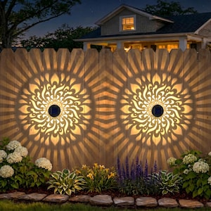 May include: Two solar-powered decorative lights mounted on a wooden fence, casting intricate floral patterns. The lights are circular with a black solar panel in the center. The fence is surrounded by lush greenery and flowers, creating a warm, inviting ambiance.