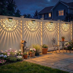 May include: Outdoor scene at dusk featuring a white fence with decorative solar lights casting intricate patterns. The lights illuminate the fence and surrounding plants, including flowers in terracotta pots, creating a warm ambiance.