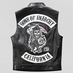 Könnte beinhalten: Schwarze Lederweste mit weißen Aufnähern mit der Aufschrift "SONS OF ANARCHY" und "CALIFORNIA". Auf dem Rücken befindet sich eine Sensemann-Grafik mit Sense und Gewehr. Die Weste hat einen Knopfverschluss am unteren Saum.