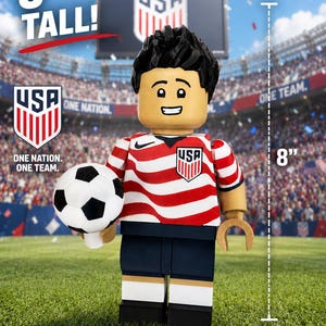 May include: A plastic figure styled like a building block toy, standing 8 inches tall. The figure wears a red and white striped jersey with a USA crest and a black Nike logo, dark blue shorts, and white socks. It holds a black and white soccer ball.