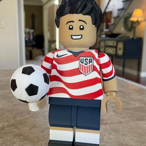 May include: A large plastic figure resembling a building block toy, featuring a person with black hair, wearing a red and white striped USA soccer jersey with a Nike logo, dark blue shorts, and white socks with black shoes, holding a soccer ball.