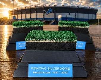 Puede incluir: Tres piezas de exhibición rectangulares con una capa de césped artificial verde sobre una base negra. Cada base tiene una placa azul con el texto PONTIAC SILVERDOME Detroit Lions 1997 - 2002. Los objetos están dispuestos en formación triangular.